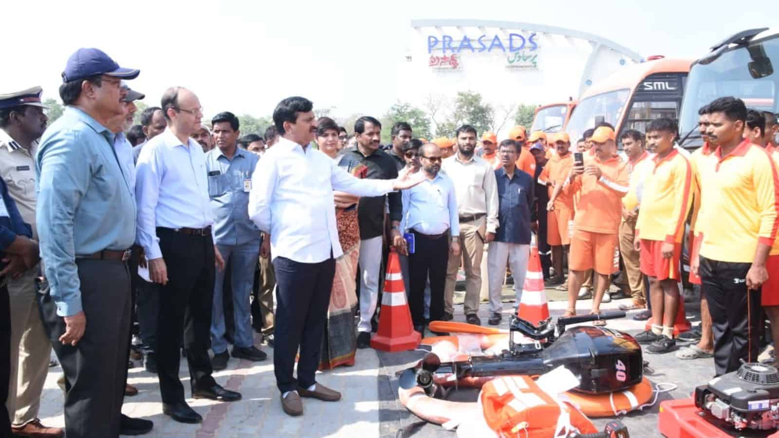 A man demonstrating safety equipment to a group of people at an outdoor event with traffic cones and a bu.
