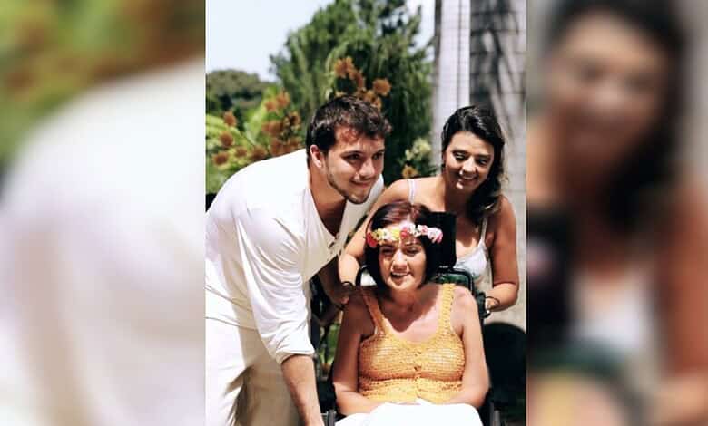 Thiago Avila stands beside his mother Teresa Regina de Avila e Silva, seated in a wheelchair, during a family gathering outdoors.