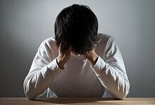 Young man with dark hair sitting at a table, head in hands, experiencing stress or anxiety.