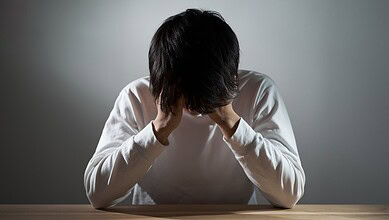 Young man with dark hair sitting at a table, head in hands, experiencing stress or anxiety.