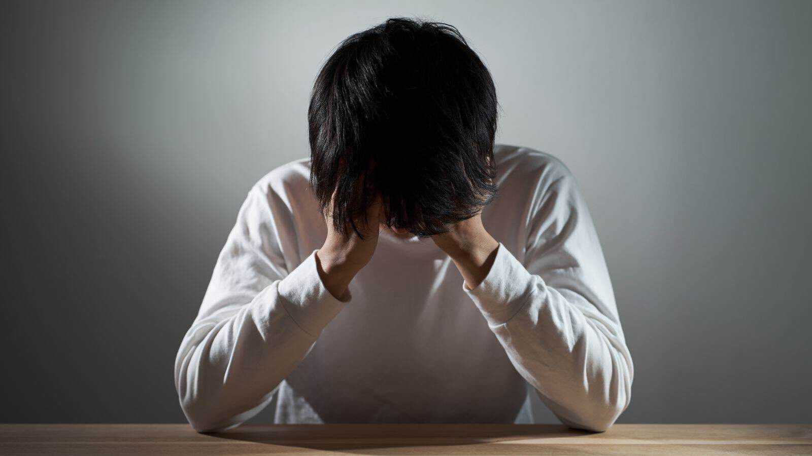 Young man with dark hair sitting at a table, head in hands, experiencing stress or anxiety.
