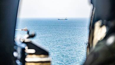 View of a cargo ship sailing across the ocean, seen from the window of a vessel, with a smaller boat near.