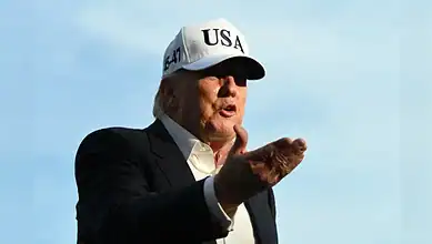 Trump in a "USA" cap, speaking outdoors with a blue sky, making a gesture during a public appearance.