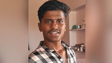 A young man smiling indoors, wearing a checkered shirt, with shelves and household items in the backgroun.