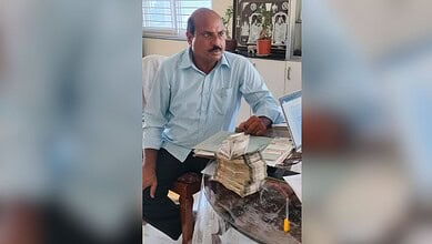 Man with a mustache sitting at a desk with stacks of cash and a laptop in an office.