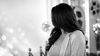 South Indian actress with long dark hair in a grey blazer, facing away in a studio setting.
