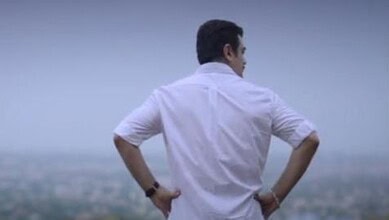 Man in white shirt with hands on hips looking at cityscape from a hilltop.