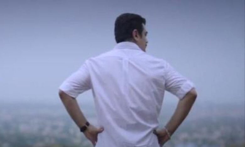 Man in white shirt with hands on hips looking at cityscape from a hilltop.