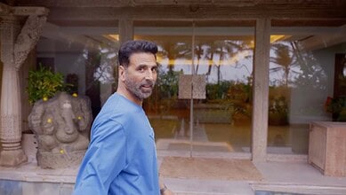 Man in blue shirt standing outdoors near a modern building with glass doors and tropical plants.