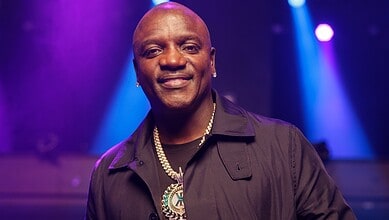 A smiling man in a black shirt with a gold chain and medallion, standing in a vibrant, colourful event se.
