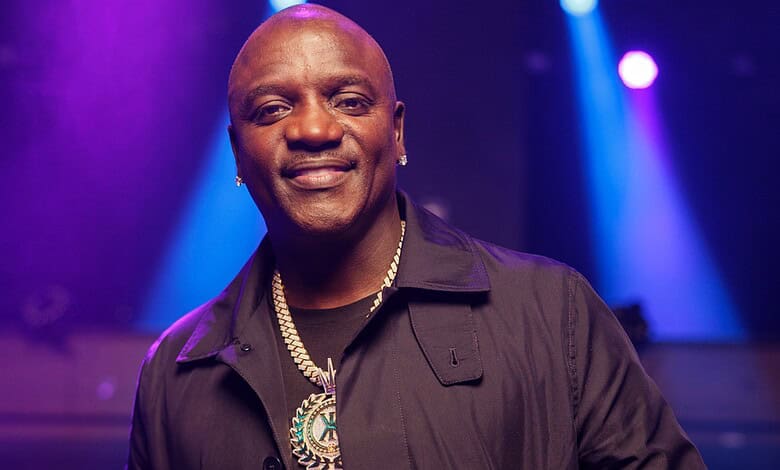 A smiling man in a black shirt with a gold chain and medallion, standing in a vibrant, colourful event se.