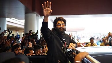 Man with glasses and beard waving to crowd at a public event, surrounded by photographers and attendees.