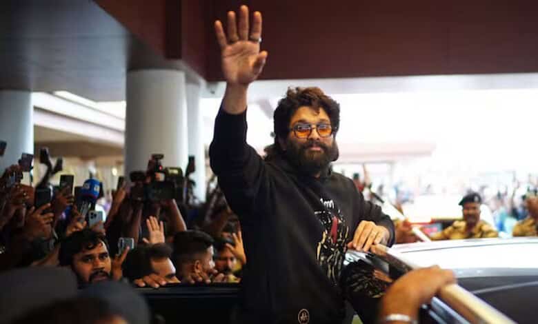 Man with glasses and beard waving to crowd at a public event, surrounded by photographers and attendees.