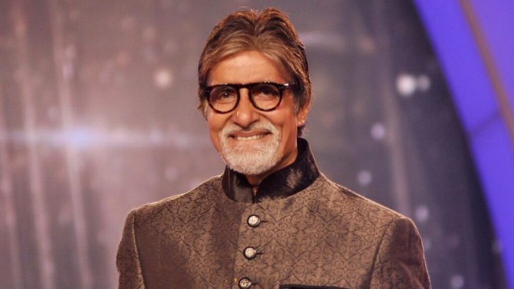 Amitabh Bachchan smiling in a formal traditional outfit, wearing glasses, on a stage with a blurred backg.