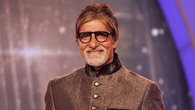 Amitabh Bachchan smiling in a formal traditional outfit, wearing glasses, on a stage with a blurred backg.