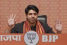 BJP leader addressing media with hands raised, seated at a table with BJP and lotus symbols in the backgr.