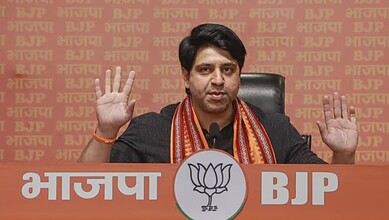 BJP leader addressing media with hands raised, seated at a table with BJP and lotus symbols in the backgr.