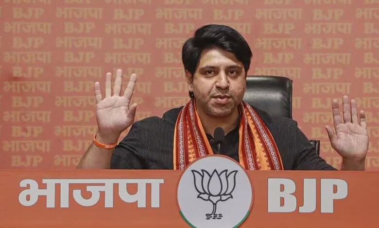 BJP leader addressing media with hands raised, seated at a table with BJP and lotus symbols in the backgr.