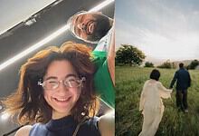 Smiling woman with glasses and curly hair capturing a selfie indoors, with a man in the background. Brigh.