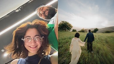 Smiling woman with glasses and curly hair capturing a selfie indoors, with a man in the background. Brigh.