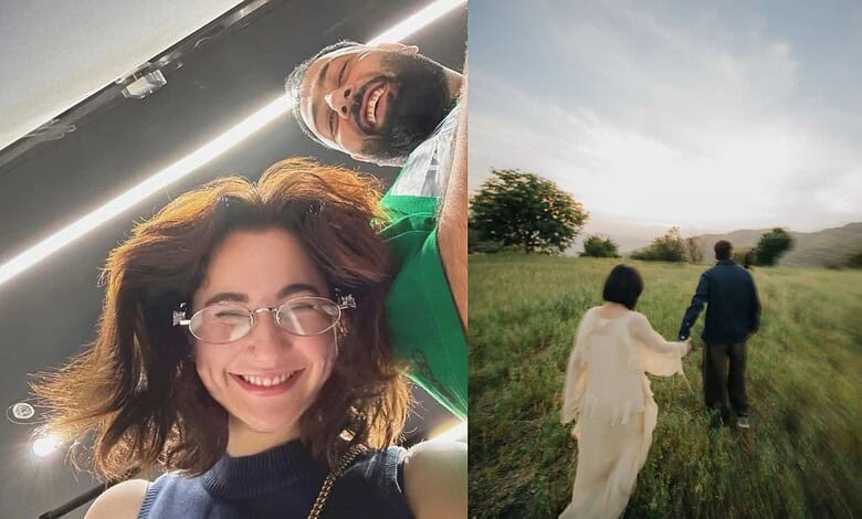 Smiling woman with glasses and curly hair capturing a selfie indoors, with a man in the background. Brigh.