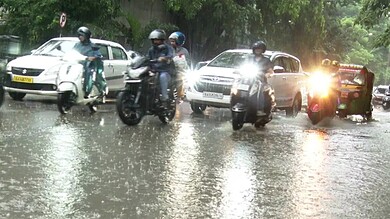 Flooded street with motorcycles and cars during heavy rain, with police vehicle in the background.
