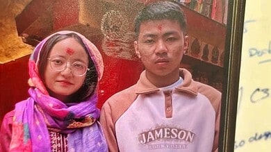 A young boy and woman wearing traditional attire, standing indoors with a red background and bookshelves.