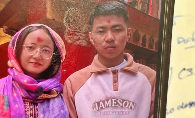 A young boy and woman wearing traditional attire, standing indoors with a red background and bookshelves.