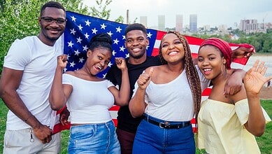 A woman and four young adults holding an American flag outdoors, celebrating diversity and unity.