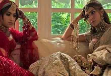 A woman in traditional red bridal attire and another in beige bridal dress sitting indoors with a window.