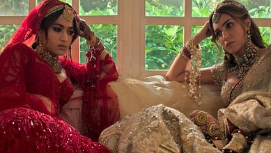 A woman in traditional red bridal attire and another in beige bridal dress sitting indoors with a window.