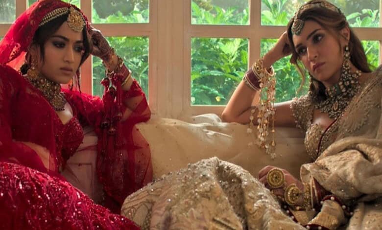 A woman in traditional red bridal attire and another in beige bridal dress sitting indoors with a window.