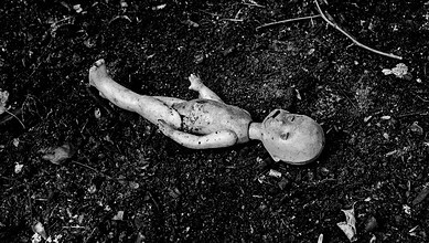 A baby doll lying on dark, dirt-covered ground with debris scattered around.