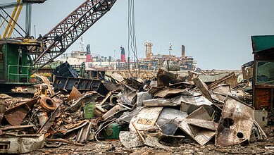 Heap of discarded metal and construction waste in an open area with cranes in the background.