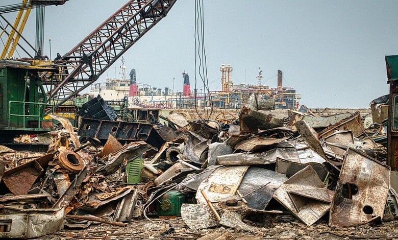 Heap of discarded metal and construction waste in an open area with cranes in the background.
