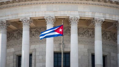 Cuban flag waving in front of a government building with classical columns and ornate architecture.