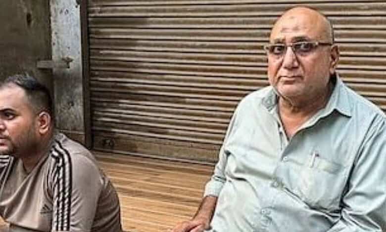 A man with glasses and a bald head sitting outdoors next to another man with short hair, in front of a cl.