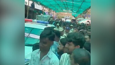 Crowd of people gathered under a green canopy in an outdoor market or street scene in India.