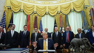 President Trump in the Oval Office with officials, with American flags and gold curtains in the backdrop.