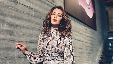 Young woman with wavy brown hair in a snake print dress standing against a concrete wall.
