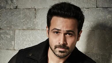 A man with dark hair and beard posing against a stone wall background.