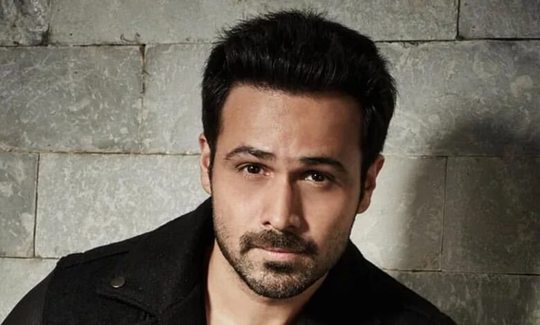 A man with dark hair and beard posing against a stone wall background.