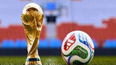 Golden FIFA World Cup trophy and a colourful football on a stadium pitch.