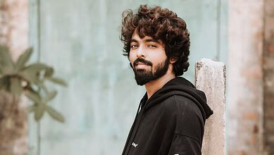 Young man with curly hair and beard leaning against a wall outdoors.