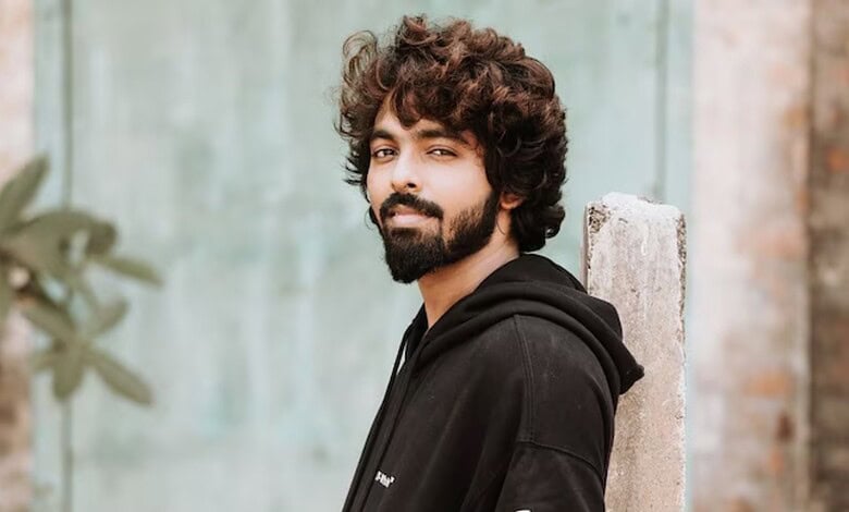 Young man with curly hair and beard leaning against a wall outdoors.