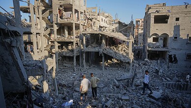 Destruction in a war-torn urban area with damaged buildings and debris, showing civilians walking through.