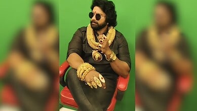 Man wearing sunglasses and traditional jewelry, sitting on a red chair against a green background.