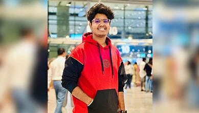 Young man with glasses in a red and black hoodie smiling at an airport terminal.