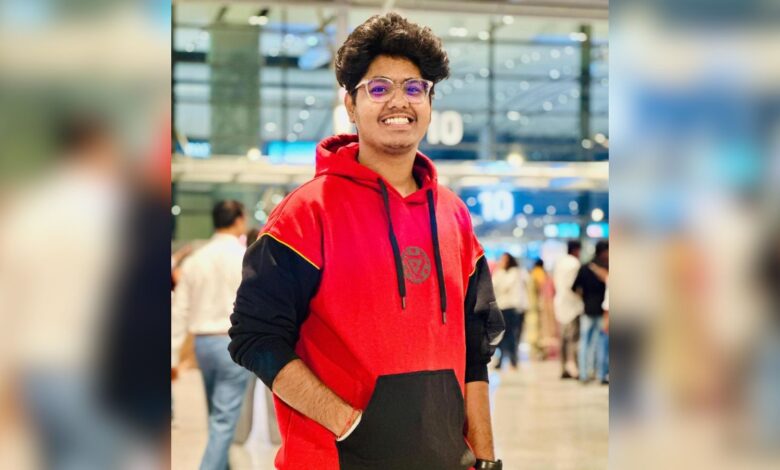 Young man with glasses in a red and black hoodie smiling at an airport terminal.