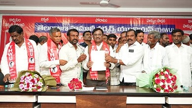 Telangana government officials and representatives at a formal event with flower bouquets and banners.